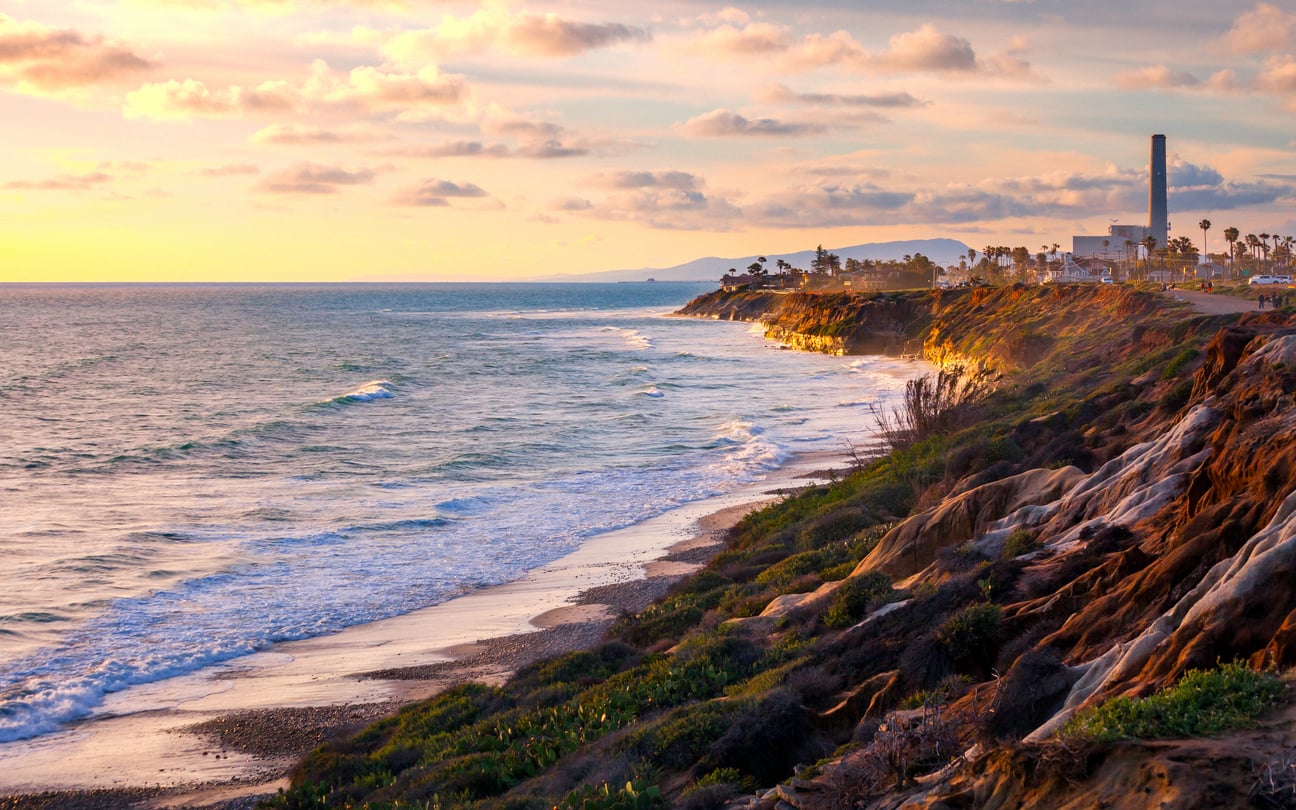 Sunny coastal scene at sunset with waves crashing against rocky cliffs, a sandy beach below, palm trees swaying gently, and a distant building with a tall smokestack—this breathtaking vista easily ranks among the top 20 travel destinations for those seeking natural beauty and tranquility.