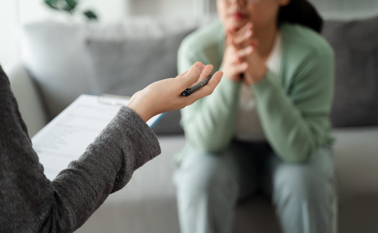A person holding a clipboard and pen talks to another person sitting on a couch, who is listening with hands clasped under their chin. The setting appears to be a casual indoor meeting or therapy session, where they might be discussing Baby Boomer beliefs and how these perspectives influence their current experiences.