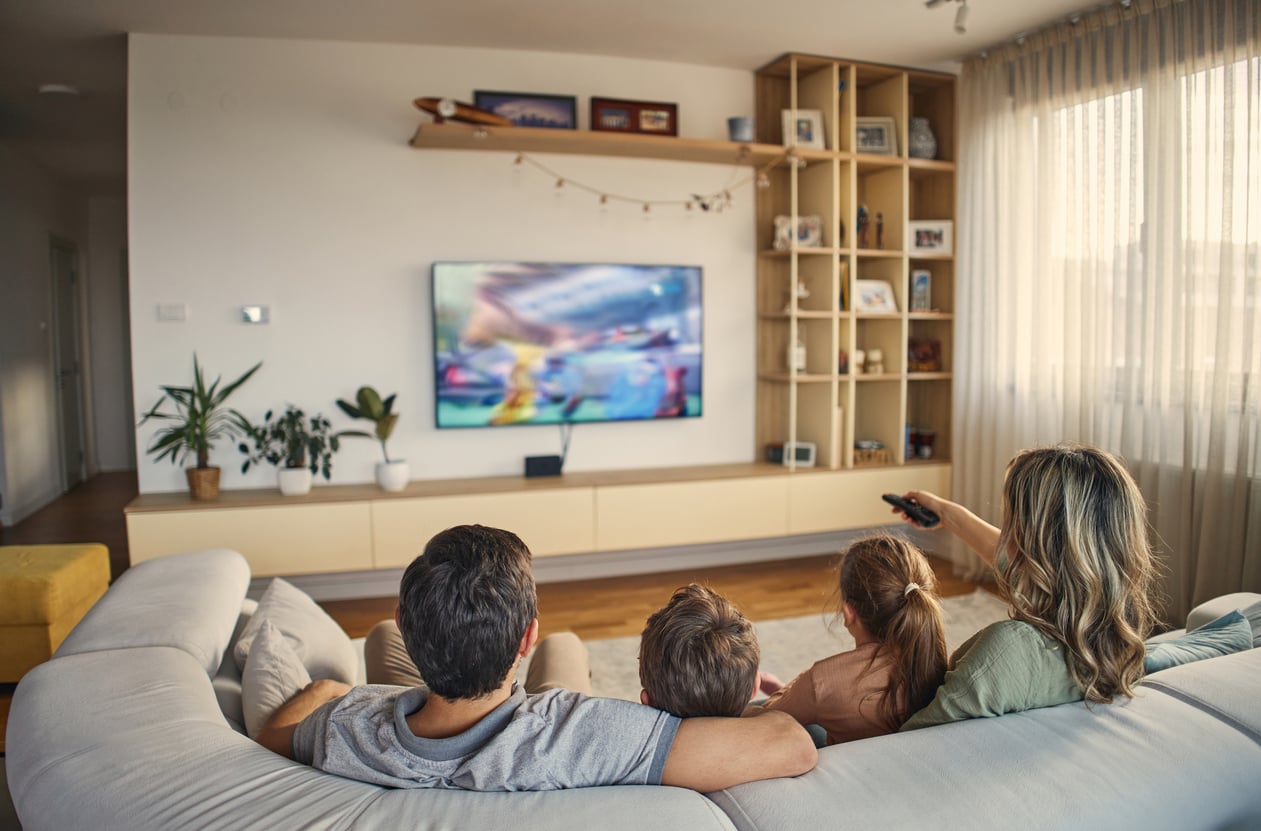 A family of three sits on a couch in a living room, watching a colorful show on a wall-mounted TV. The woman holds a remote. Shelves, plants, and a large window with curtains are visible in the cozy space.