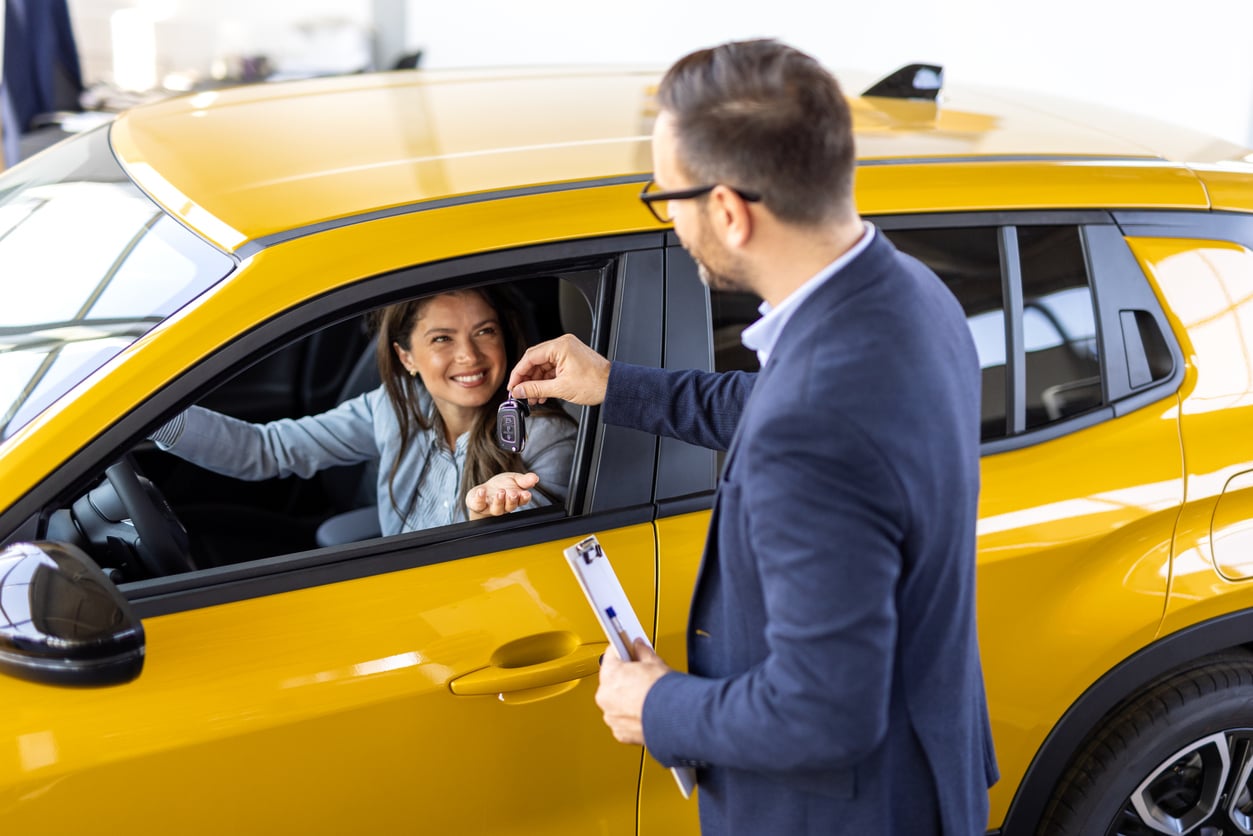 A man hands car keys to a smiling woman sitting in the driver’s seat of a yellow car, suggesting a car purchase or rental and highlighting ways to manage vacation costs.