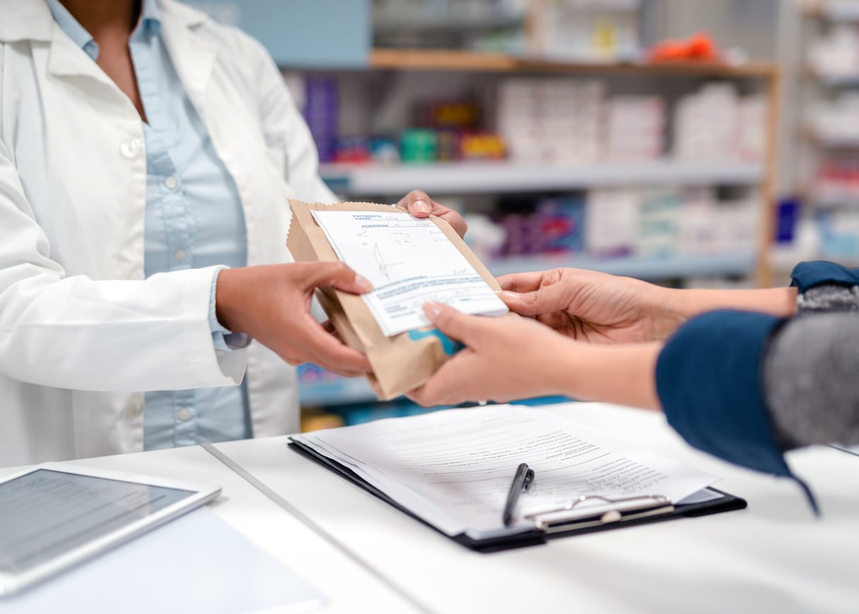 A pharmacist hands a paper bag with a prescription label to a customer across a pharmacy counter, ensuring they receive only what’s necessary and not waste of money products. A clipboard with papers and a tablet are on the counter. Shelves with medicine are in the background.