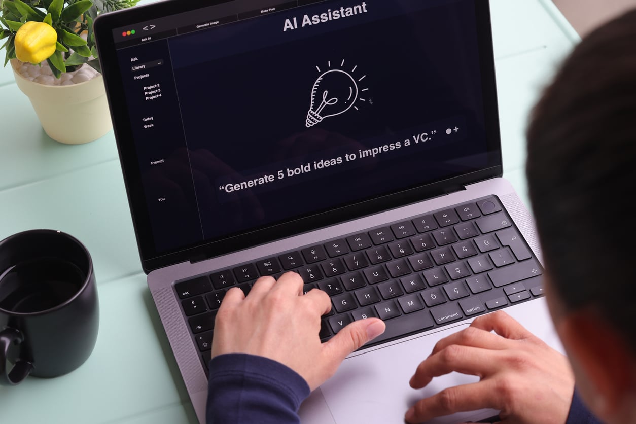 A person types on a laptop displaying an AI assistant screen with a lightbulb icon and the prompt: “Generate 5 bold ideas to impress a VC.” A black mug and a small potted plant are on the desk.