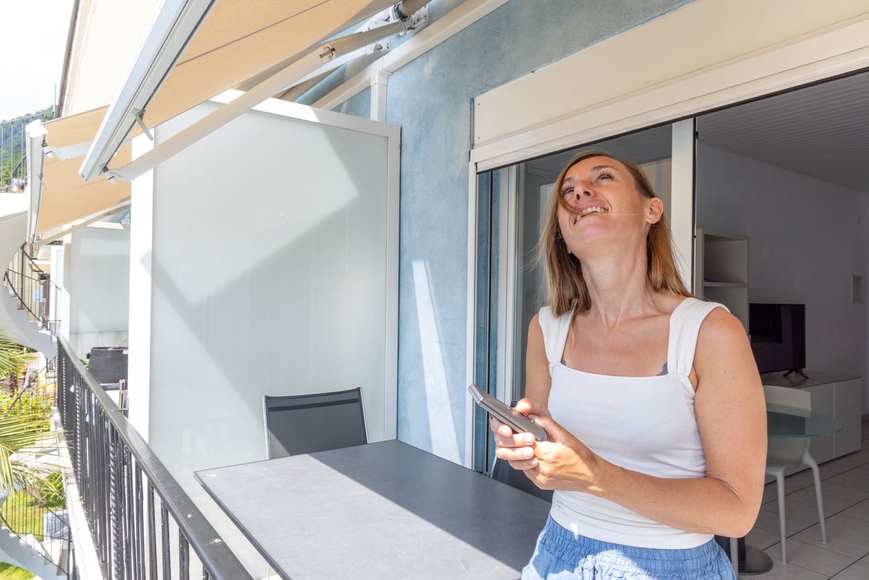 A woman stands on a balcony holding a smartphone, smiling and looking up. She wears a white tank top and blue skirt, next to a black table featuring outdoor home products. The scene appears bright and sunny.