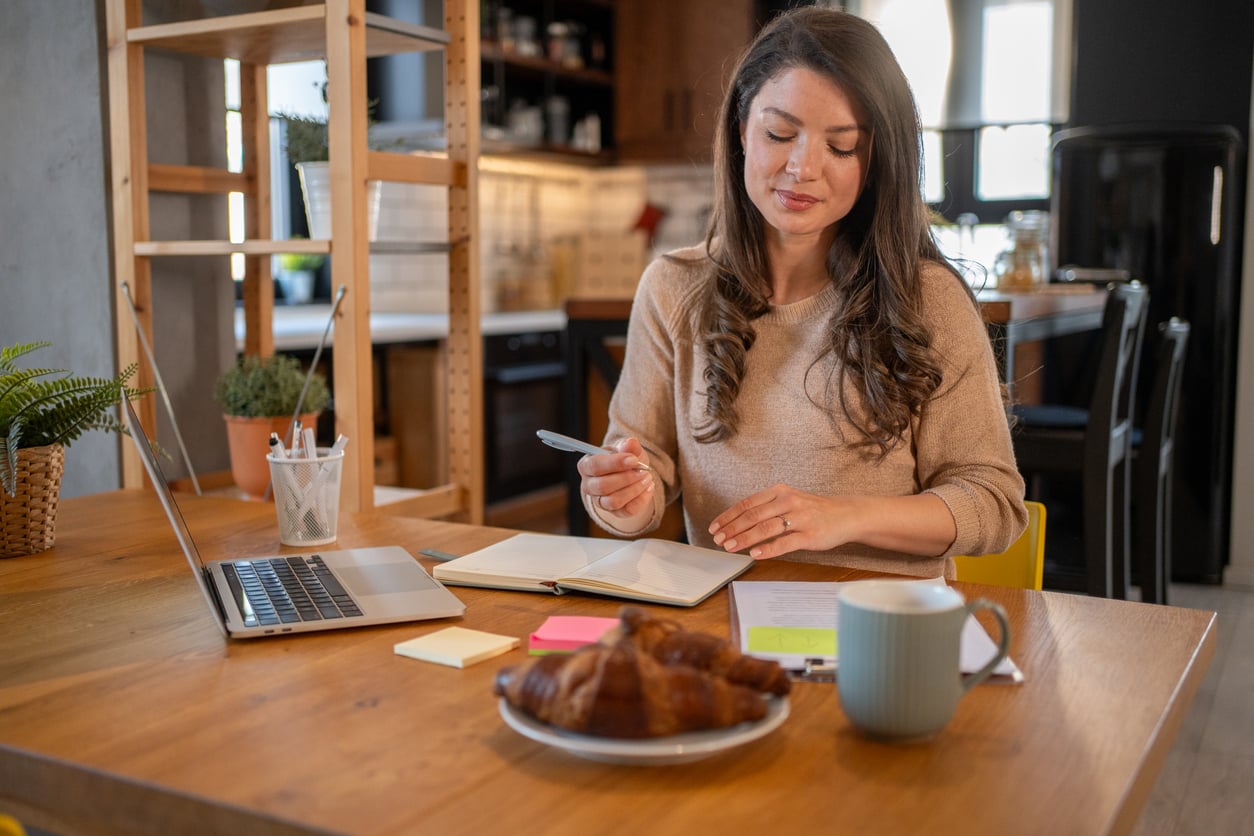 A woman sits at a wooden table with a laptop, notebook, pen, and sticky notes. A croissant and a mug are in the foreground. She appears to be working or studying in a cozy kitchen setting.