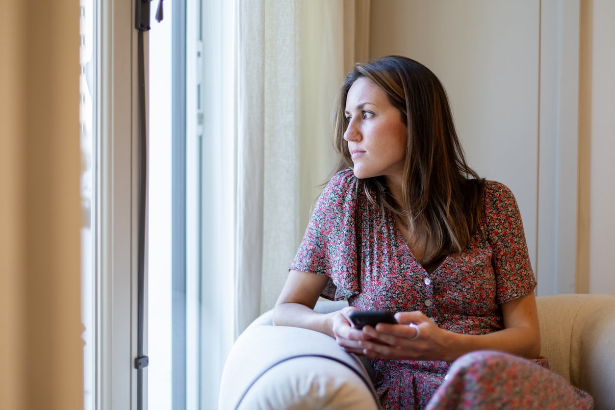 A woman in a floral dress sits in an armchair, holding a smartphone and looking thoughtfully out a window with light curtains, perhaps reflecting on her money saving habits.