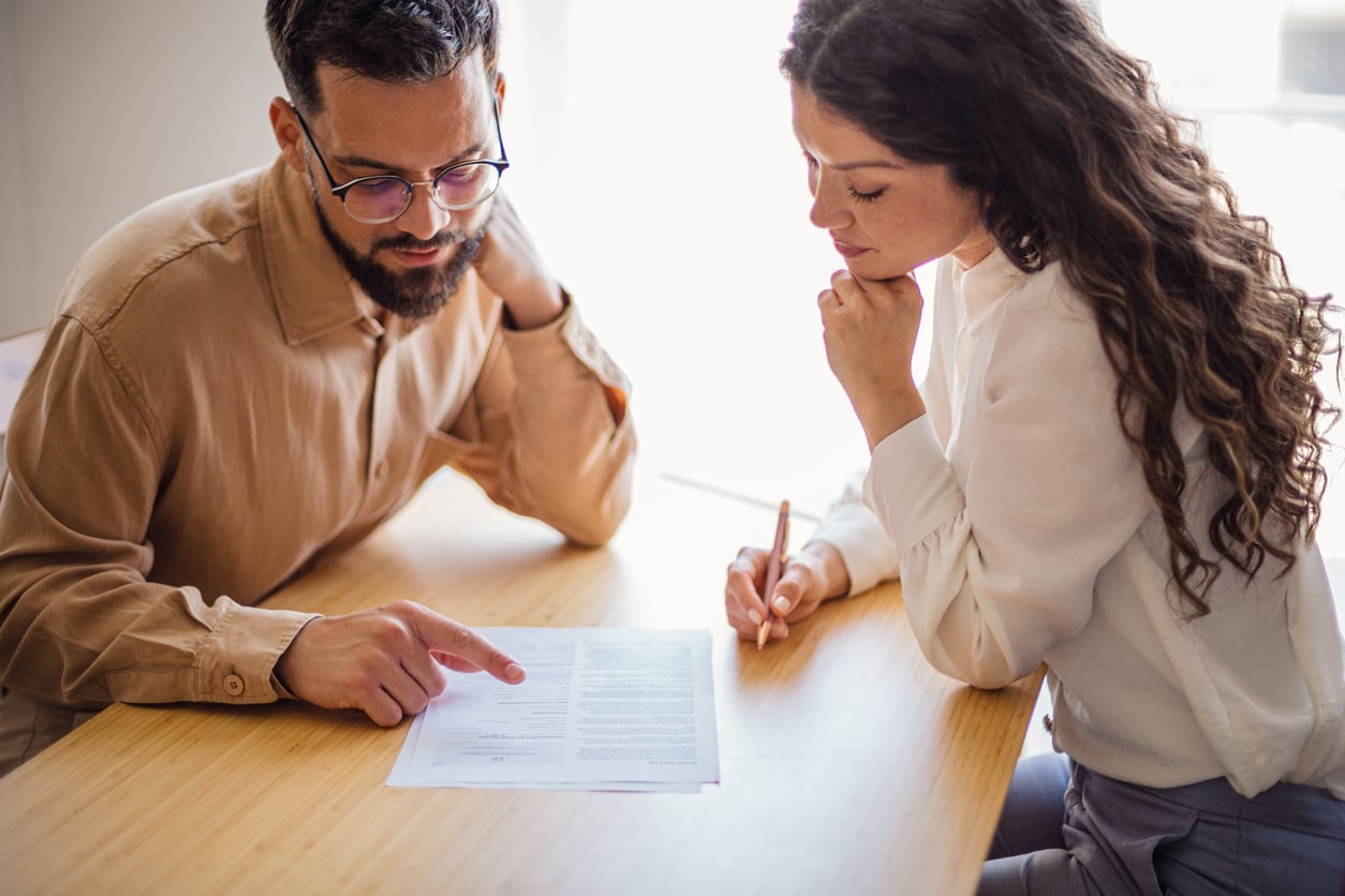 Two people sit at a table reviewing a document together. One person points to the paper while the other holds a pen, appearing thoughtful and focused on the content in front of them as they discuss effective money saving habits.