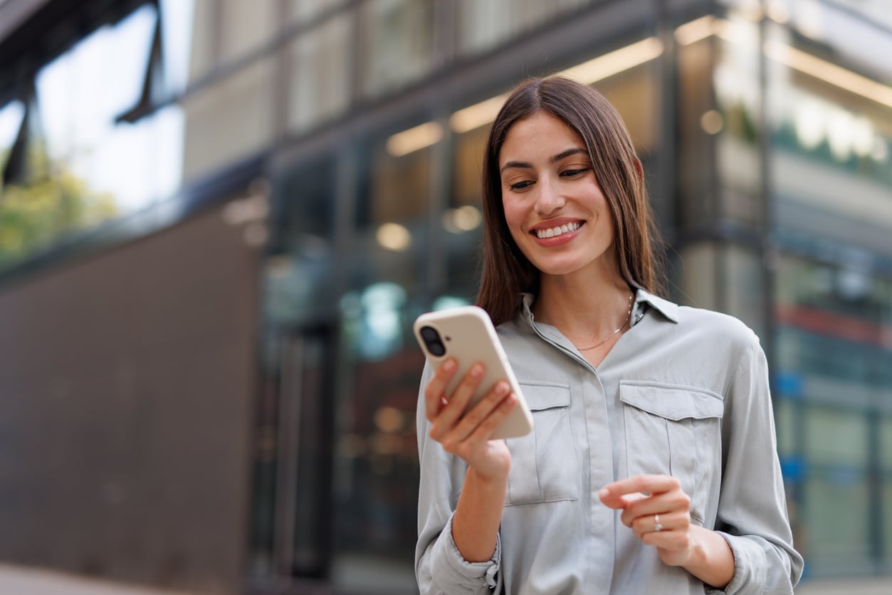 A woman with long brown hair smiles while looking at her smartphone outside a modern glass building. She wears a light gray button-up shirt and appears happy and engaged with her device, perhaps checking on her monthly bills with ease.