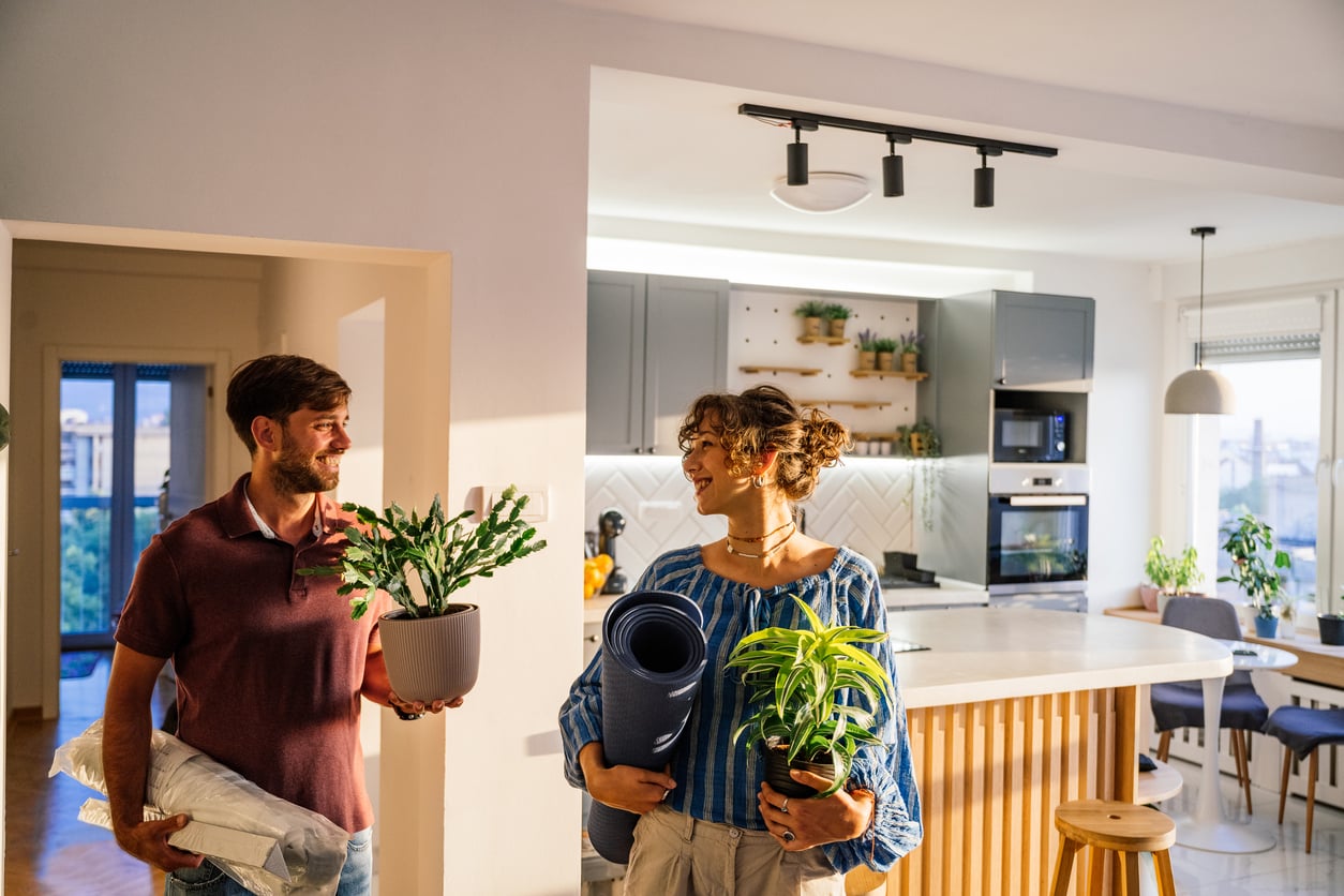A smiling man and woman stand in a modern kitchen, holding houseplants and household items, looking at each other happily. The kitchen has wooden accents and is well-lit with natural and artificial light, reflecting classic Baby Boomer beliefs in home comfort and togetherness.