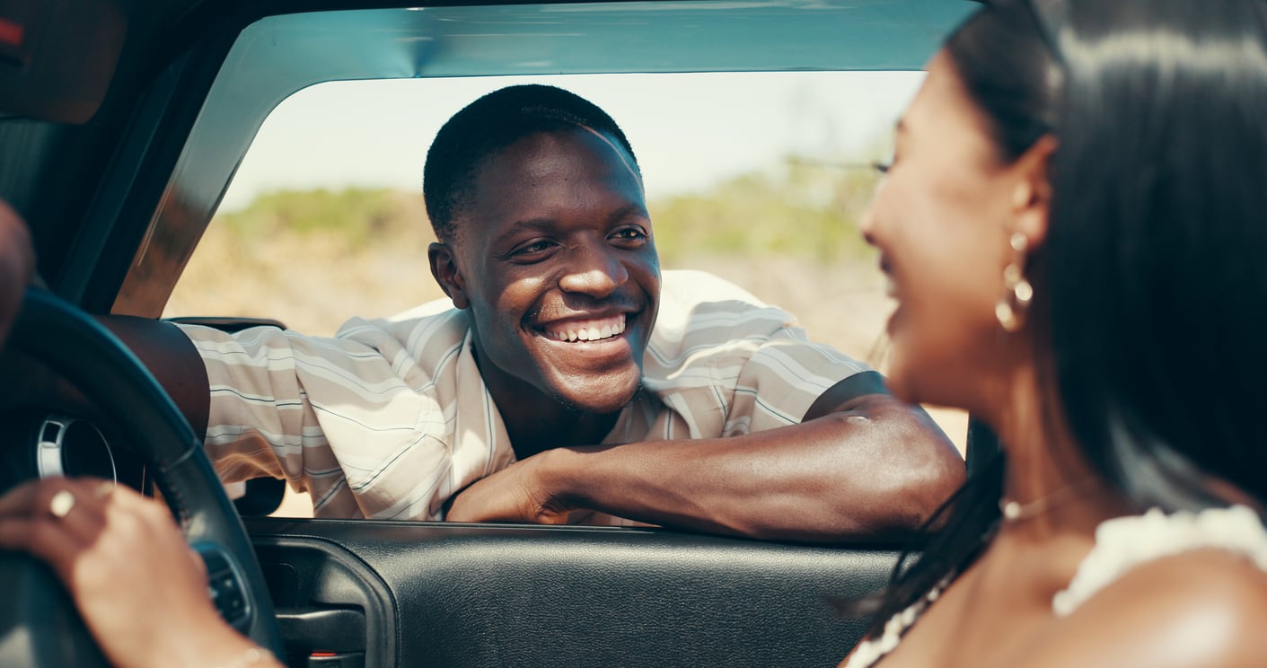 A man smiles and leans on a car window while talking to a woman in the driver's seat. They appear to be enjoying a sunny day outdoors, both wearing light, casual clothes—perhaps taking a break as they go over their road trip checklist.
