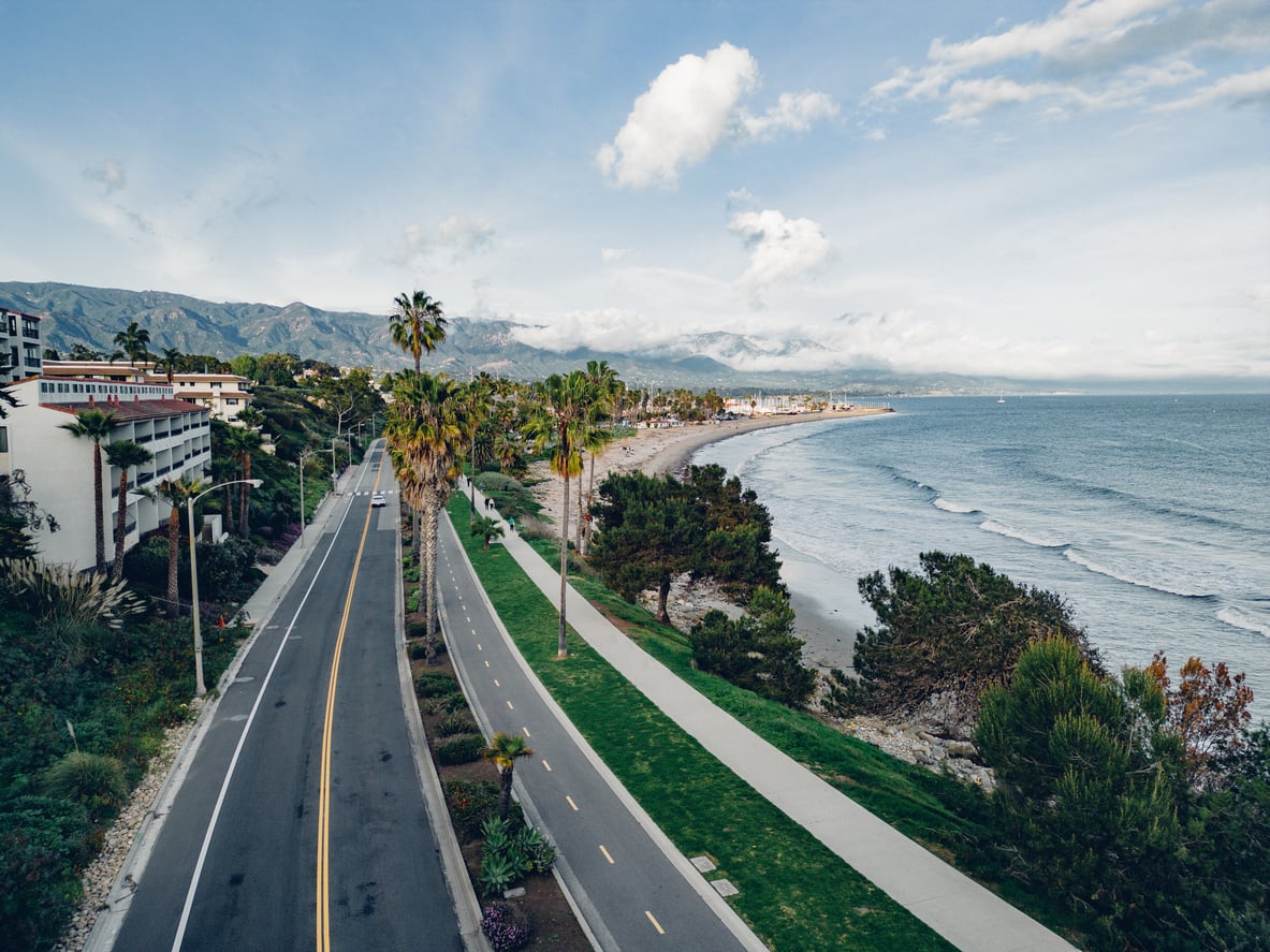A coastal road lined with palm trees runs parallel to a sandy beach and ocean waves, making it one of the most picturesque travel destinations, with mountains and buildings in the background under a partly cloudy sky.