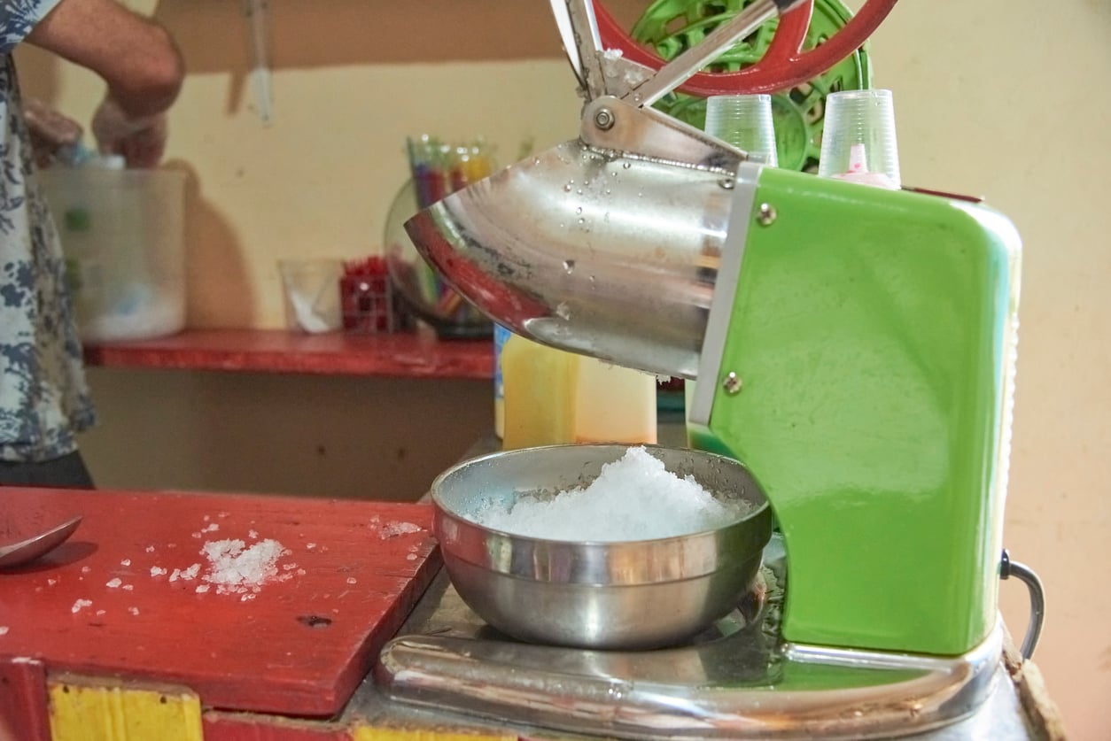 A green and silver ice-shaving machine, one of the essential kitchen gadgets, dispenses shaved ice into a metal bowl on a counter, with cups stacked nearby and a person working in the background.