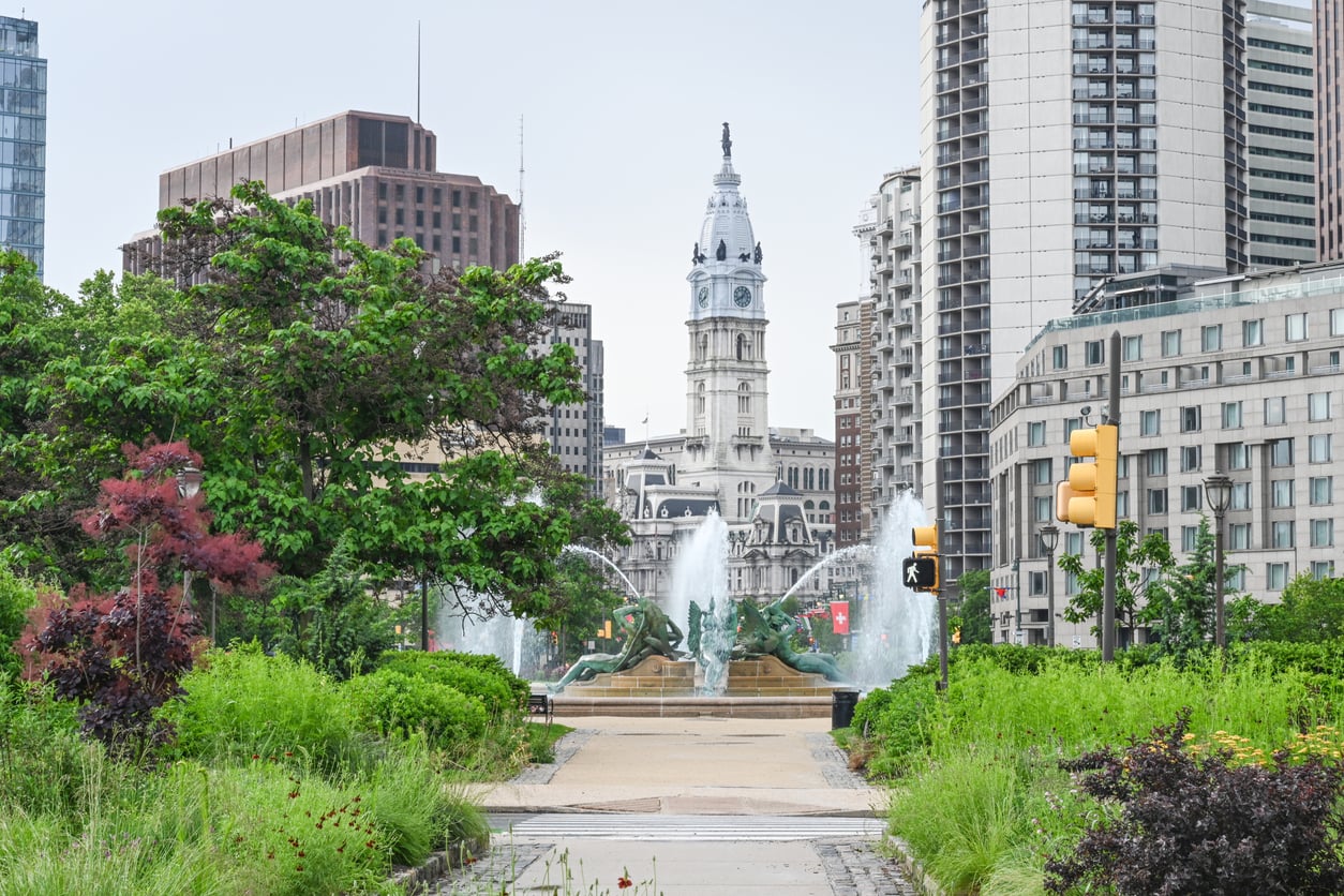 A cityscape view of Philadelphia’s City Hall tower, framed by tall buildings, green trees, fountains, and landscaped gardens in the foreground.