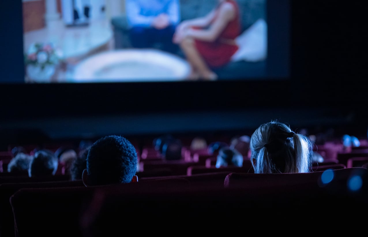People sitting in a dark movie theater, watching a film on a large screen that shows two people sitting by a round table.