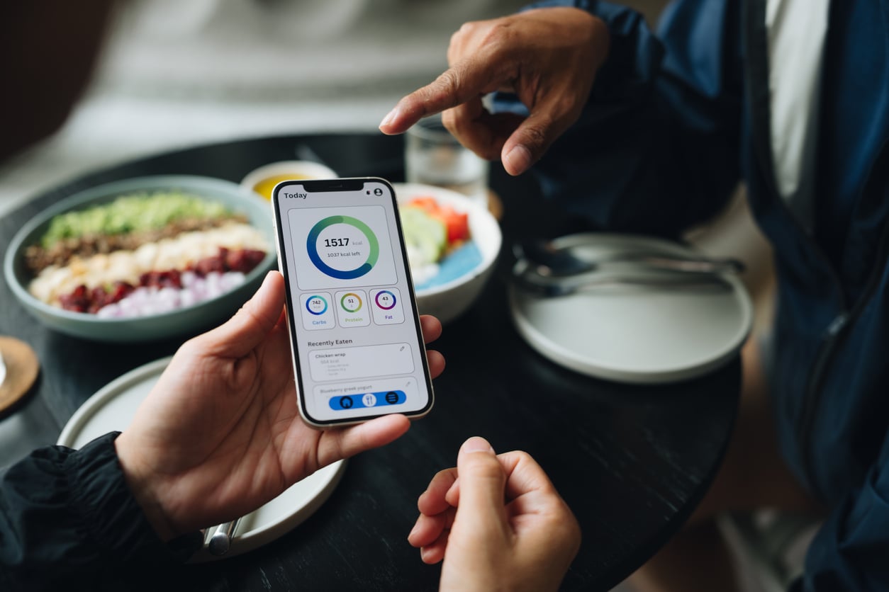 A person holds a smartphone displaying a fitness tracking app while sitting at a table with bowls of food, as another person points at the phone, perhaps discussing how staying healthy could help manage monthly bills.