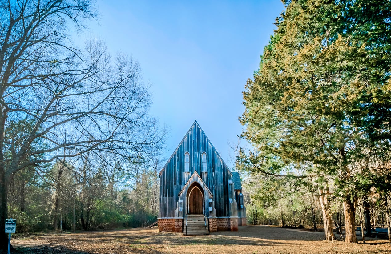 A small, rustic wooden church with a peaked roof stands centered in a clearing surrounded by trees, reminiscent of those found in classic American towns. A single wooden door is centered at the front, accessed by a short set of steps, with tall, narrow windows above. The sky is bright blue.