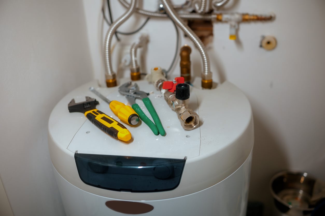 A close-up of a water heater with various products, including a wrench, screwdriver, pliers, and a yellow utility knife, placed on top. Pipes and valves are visible behind the heater.