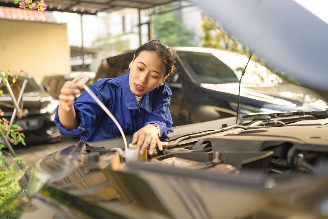 A woman in a blue jumpsuit checks a car’s oil level with a dipstick under an open hood in a sunlit garage, focused on routine maintenance that helps avoid costly monthly bills. Several vehicles are parked in the background.