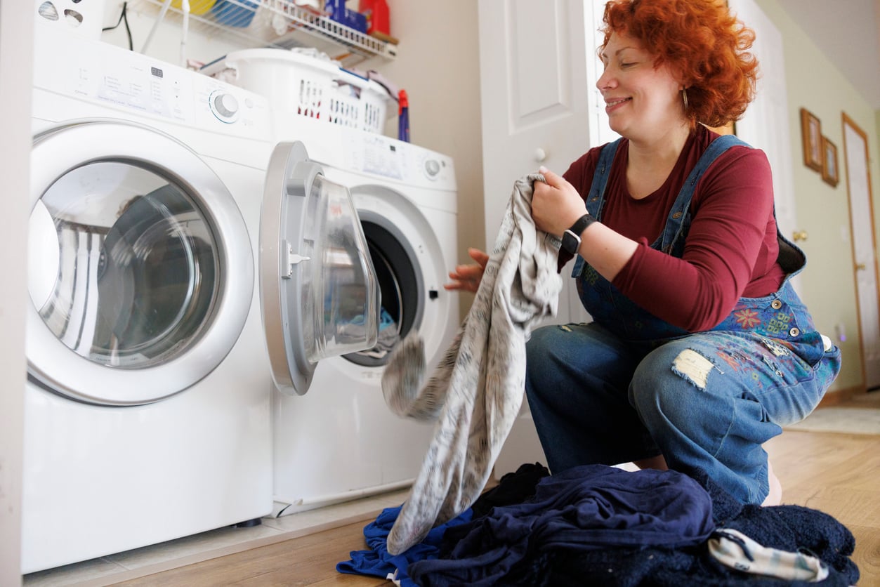 A person with curly red hair kneels in front of a washing machine, smiling as they load laundry from a pile of clothes on the floor into the open washer.