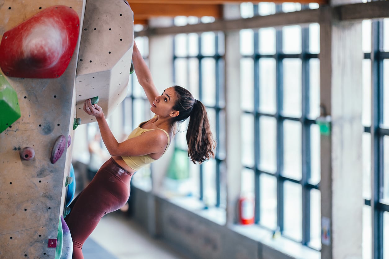 A woman in athletic wear climbs an indoor bouldering wall, gripping colorful holds—a perfect example of memberships worth paying for. Natural light streams through large windows in the background.