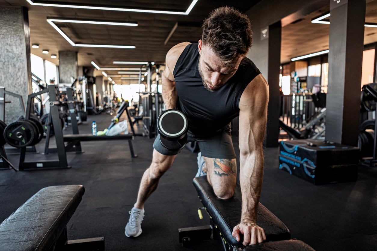 A man in athletic wear, known for his money saving habits, does a one-arm dumbbell row on a bench in a gym, surrounded by fitness equipment.