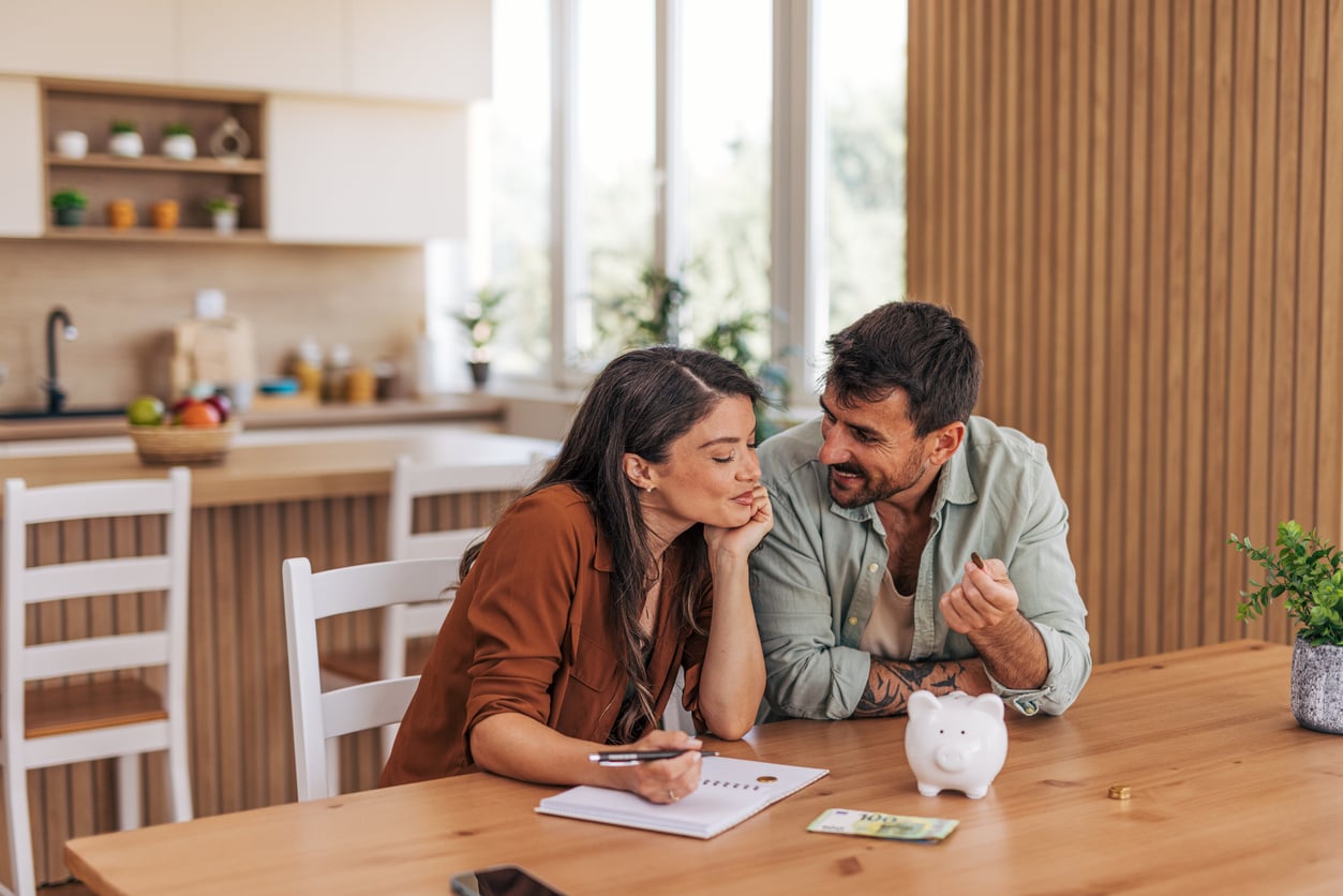 A couple sits at a kitchen table, smiling and talking. The woman writes on a notepad while the man gestures. A piggy bank and cash are on the table, suggesting a financial discussion or planning.