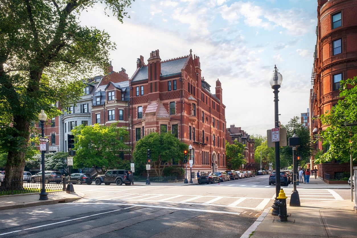 Best neighborhoods to stay in. A tree-lined city street with historic red-brick buildings, a crosswalk, streetlights, parked cars, and green trees under a partly cloudy sky.