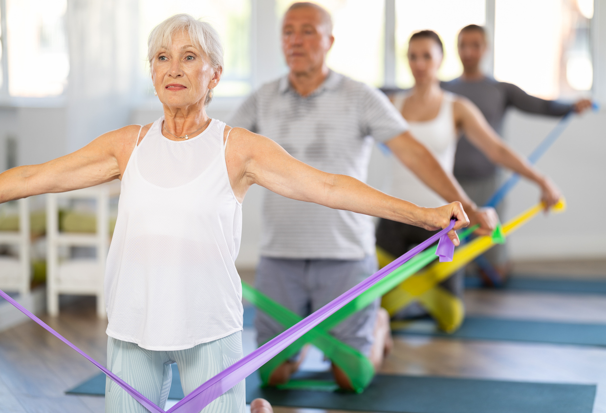 A group of older adults in athletic wear exercise indoors using colorful resistance bands, kneeling on yoga mats with arms extended to the sides. Staying active and healthy can help save money on future healthcare costs. The background is bright and softly blurred.
