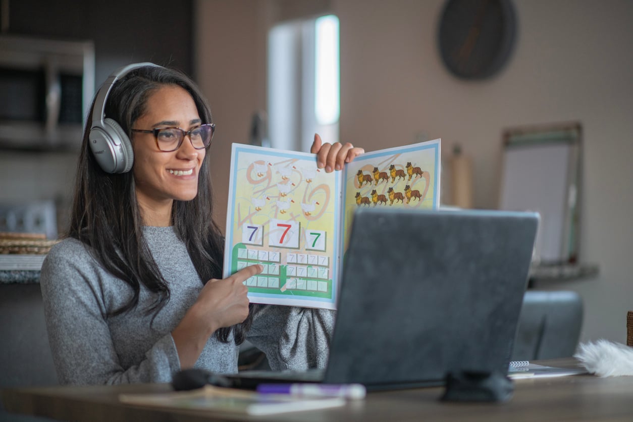 A woman wearing headphones sits at a table, smiling and holding up a children's picture book to a laptop, likely teaching or reading during a virtual lesson.