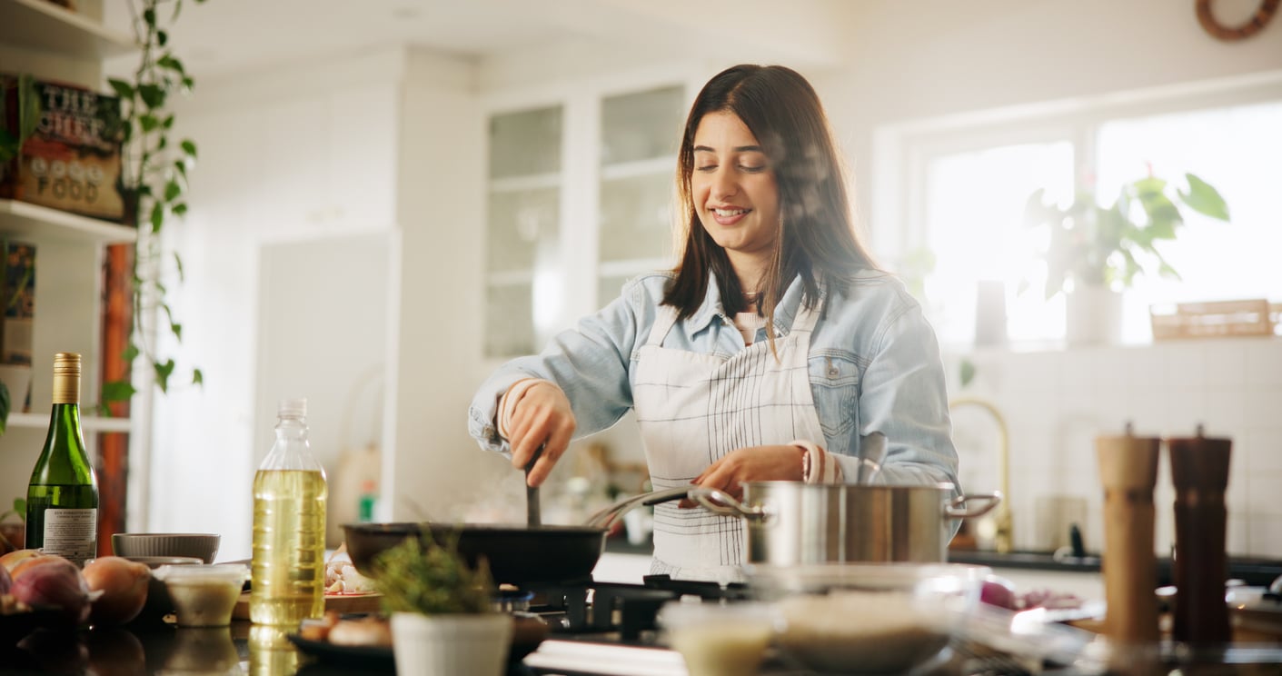 A woman wearing an apron smiles while cooking at a stove in a bright, modern kitchen. Cooking ingredients, utensils, and bottles are arranged on the counter around her.