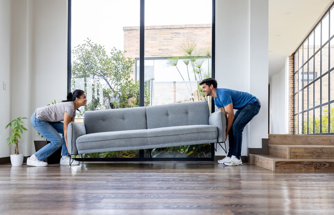 A woman and a man smile as they lift a gray sofa together in a bright living room with large windows and wooden floors, reminding us of the warmth found in cherished spaces—even as some products people no longer buy quietly fade from our homes.