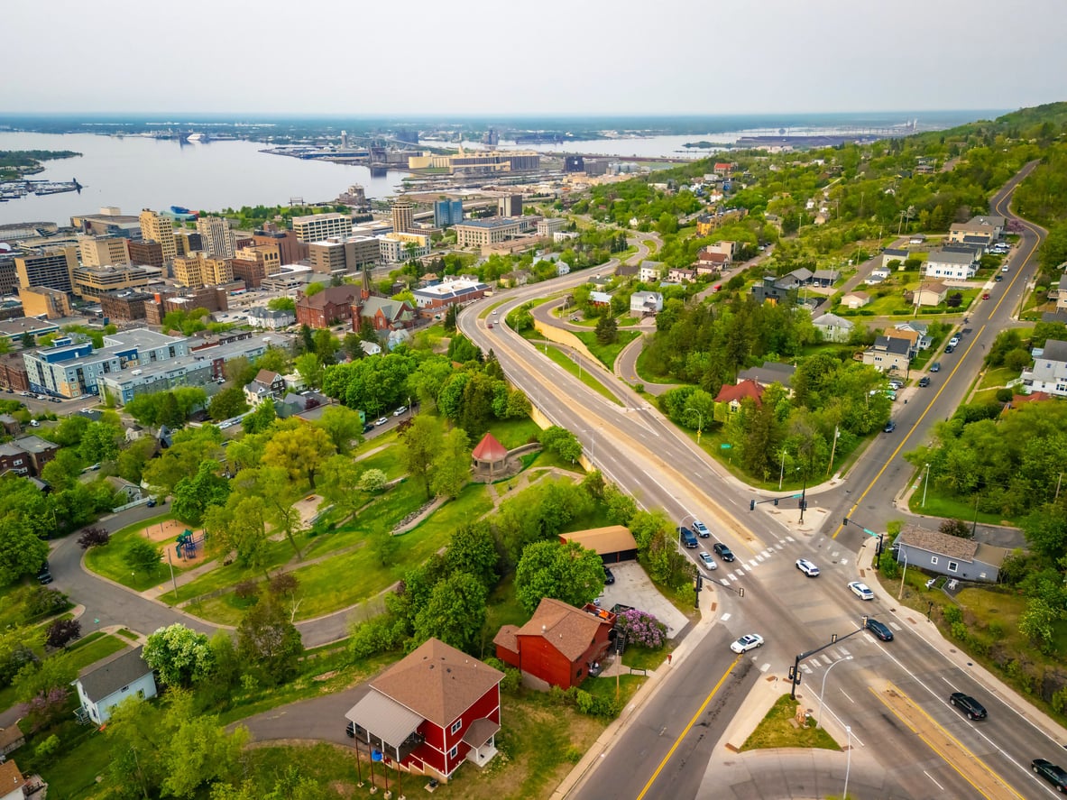 Aerial view of a city with green trees, roads, parked and moving cars, houses, park spaces, and a waterfront with buildings visible in the background—one of the 20 travel destinations known for its scenic urban landscapes and vibrant atmosphere.