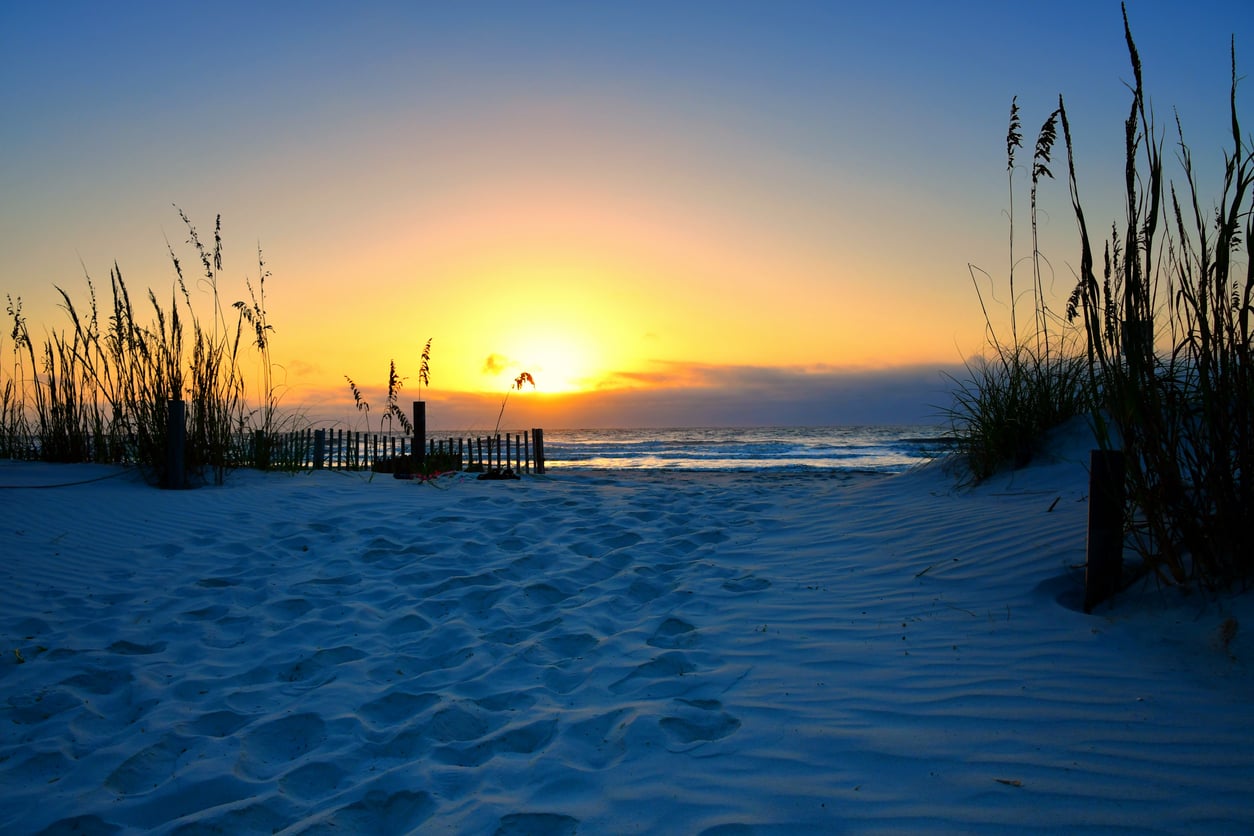 A sandy path lined with tall grass leads to the ocean, where the sun is setting on the horizon, casting a warm glow over the beach and water.