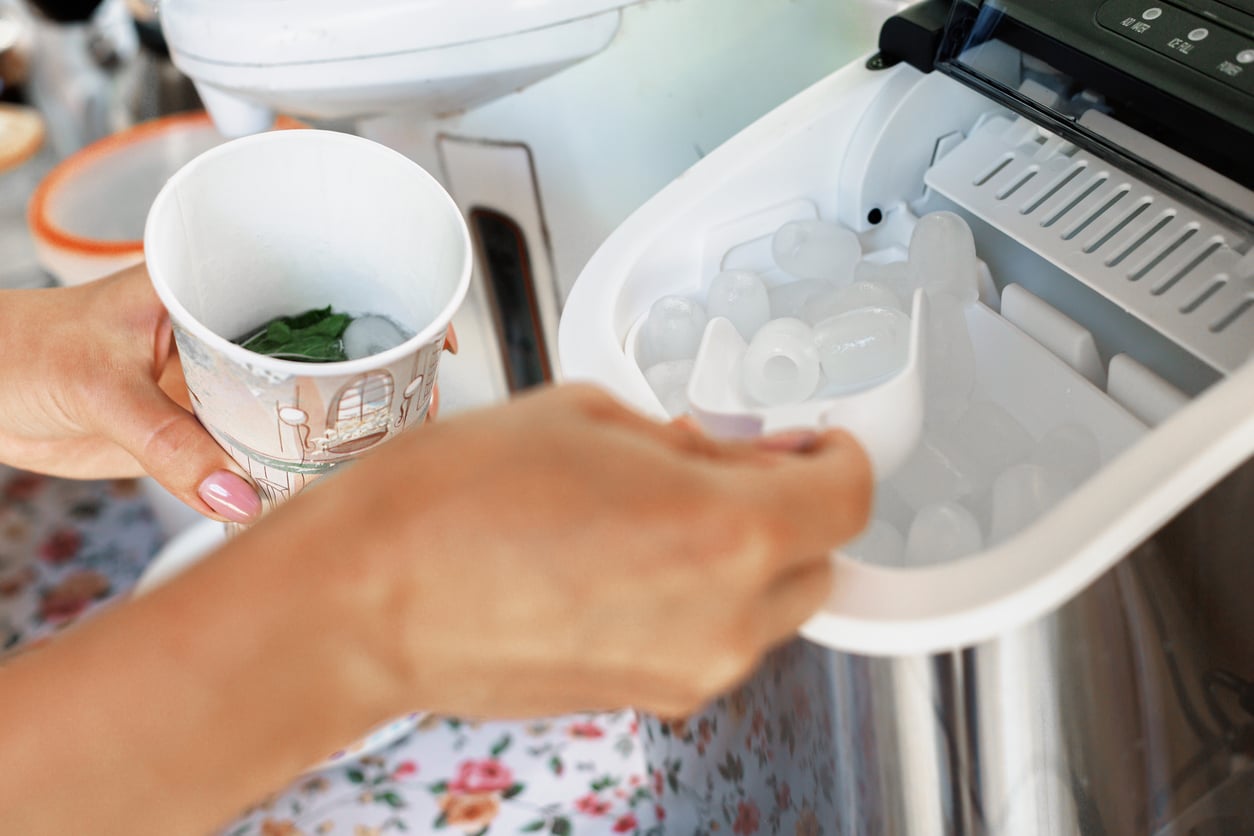 A person scoops ice cubes from an ice maker into a paper cup with mint leaves, on a floral patterned surface.
