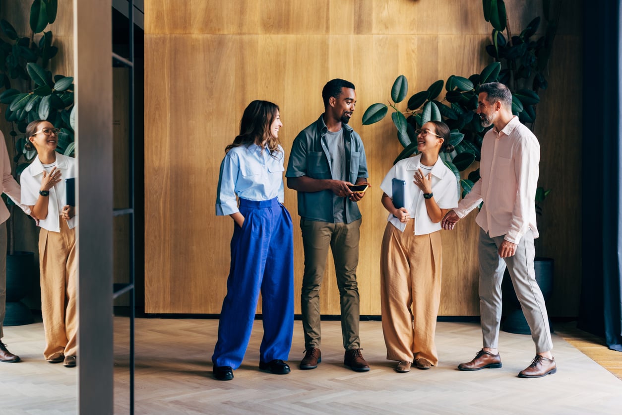 Four people stand in an office, smiling and talking. Two women and two men are casually dressed, standing near a wooden wall with plants in the background. The scene appears friendly and collaborative.