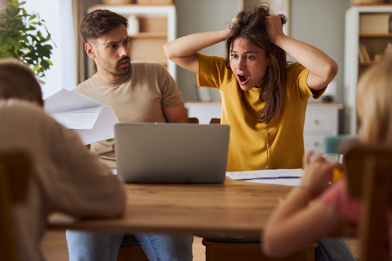 A woman sits at a table, looking stressed with her hands in her hair and mouth open. A man beside her looks concerned, holding papers. Two children are also at the table. A laptop and documents are in front of them.