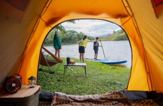 View from inside a tent showing three people by a lakeshore; one is paddleboarding, another stands nearby, and the third is watching, with camping gear set up on the grass.