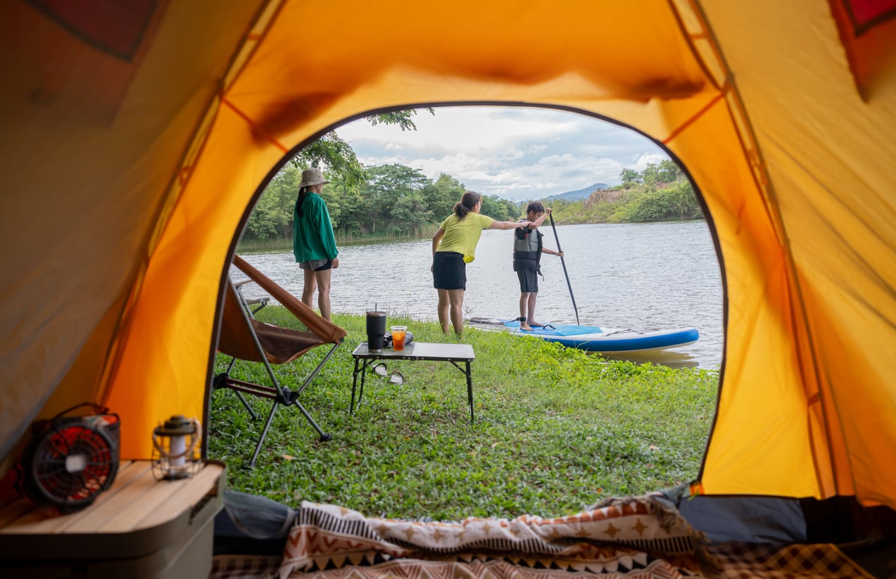 View from inside a tent showing three people by a lakeshore; one is paddleboarding, another stands nearby, and the third is watching, with camping gear set up on the grass.