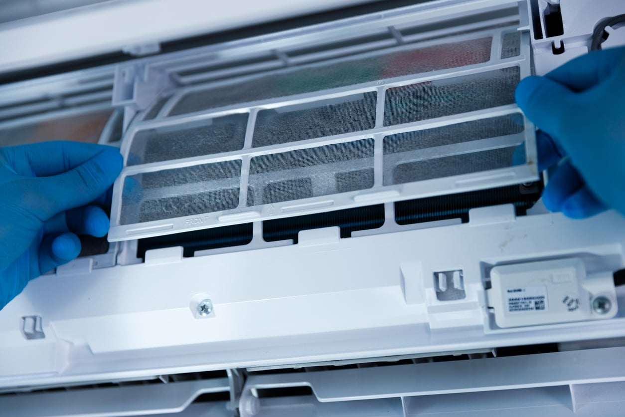 Close-up of gloved hands removing a dust filter from a white air conditioning unit for cleaning or maintenance, an important step in reducing household expenses.