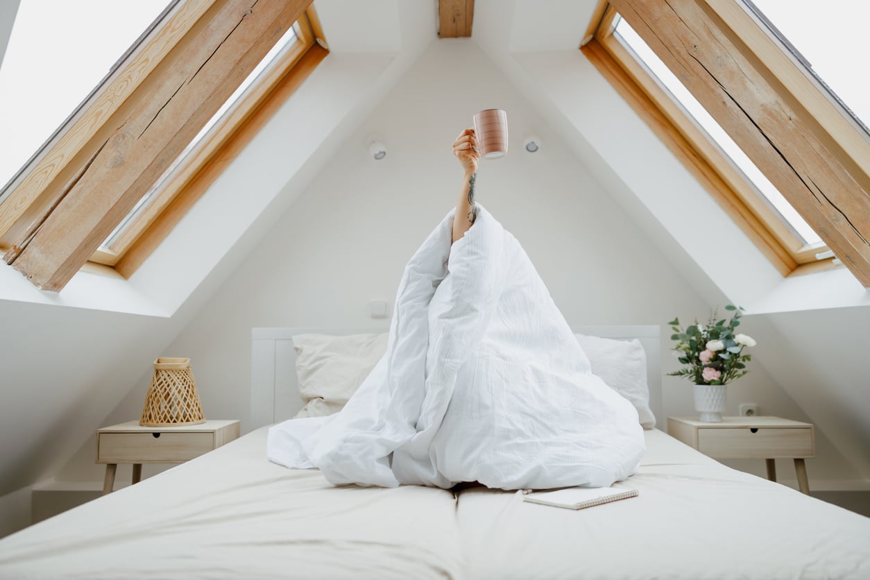 A person sits on a bed under a white blanket, raising a mug in one hand. The room has sloped ceilingsāa common example of home features homeowners no longer wantāwith skylights, wooden accents, bedside tables, flowers, and a notebook nearby.