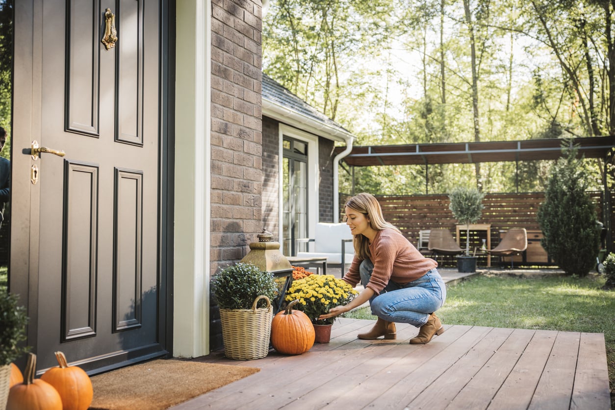 A woman crouches on a porch, arranging yellow flowers and pumpkins beside a front door. The scene features autumn decorations, outdoor home products, plants, and a backyard with outdoor seating visible in the background.