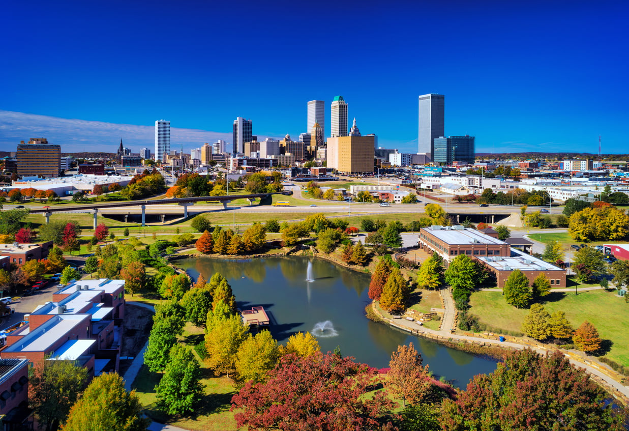 Aerial view of a travel-friendly city skyline with tall buildings, highways, and a park featuring a pond, water fountains, and colorful autumn trees under a clear blue sky.