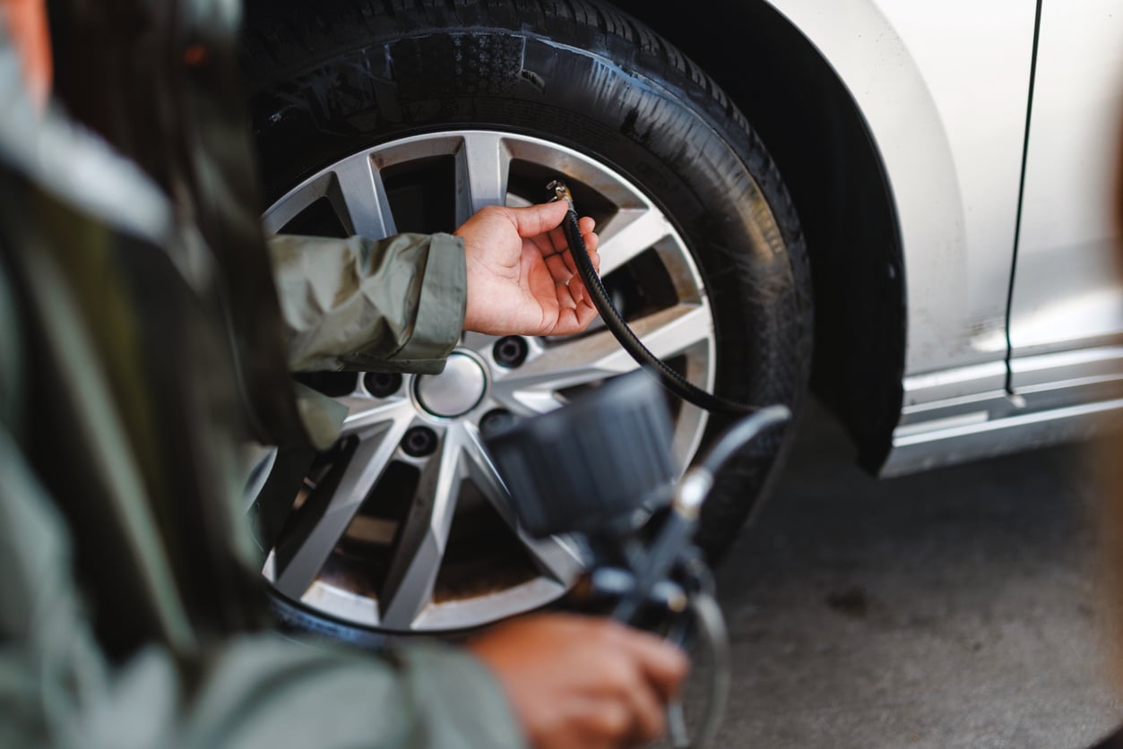 A person wearing a green jacket demonstrates car maintenance hacks by using an air pressure gauge to inflate a car tire, holding the nozzle against the valve on a silver alloy wheel.