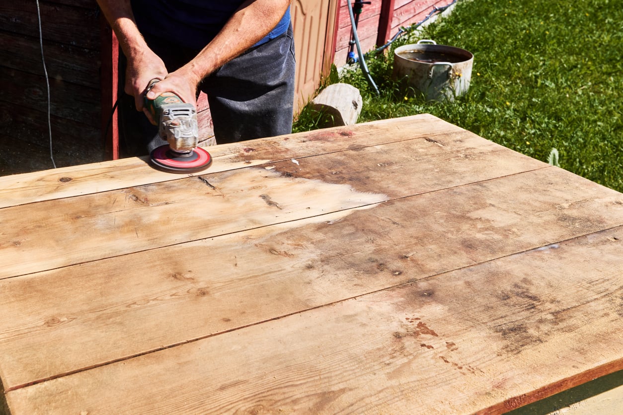 A person uses an electric sander, one of the essential products for woodworking, to smooth the surface of a large wooden table outdoors, with grass, a pot, and a red building visible in the background.