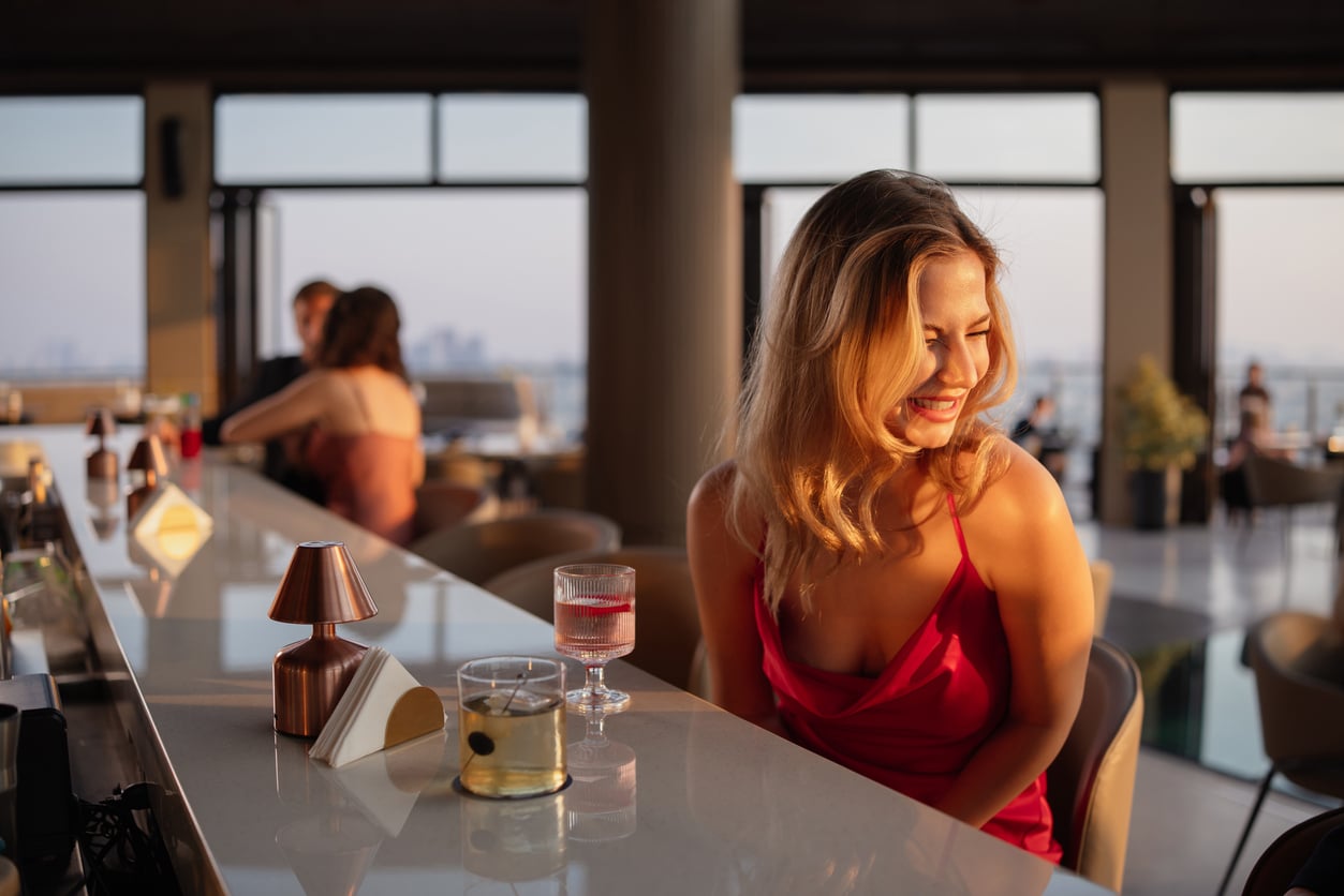 A woman in a red dress smiles at a sunlit bar, with two drinks in front of her. Other people sit in the background, and large windows reveal a cityscape outside.