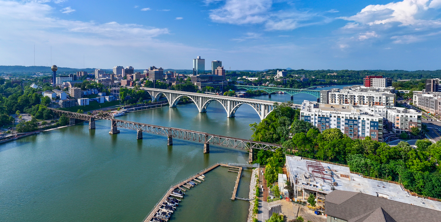 Aerial view of a city with two large bridges crossing a wide river, surrounded by buildings, greenery, and a marina with boats under a partly cloudy sky—this stunning scene easily ranks among the top 20 travel destinations for those seeking picturesque urban landscapes.