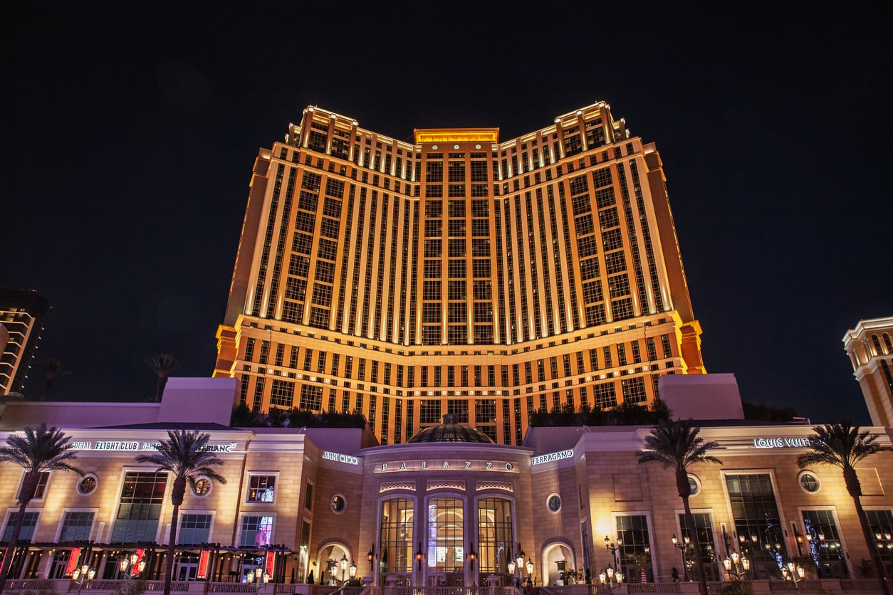 A tall, brightly lit hotel with palm trees in front, photographed at night against a dark sky. The building’s lights highlight its symmetrical architecture and grand entrance.