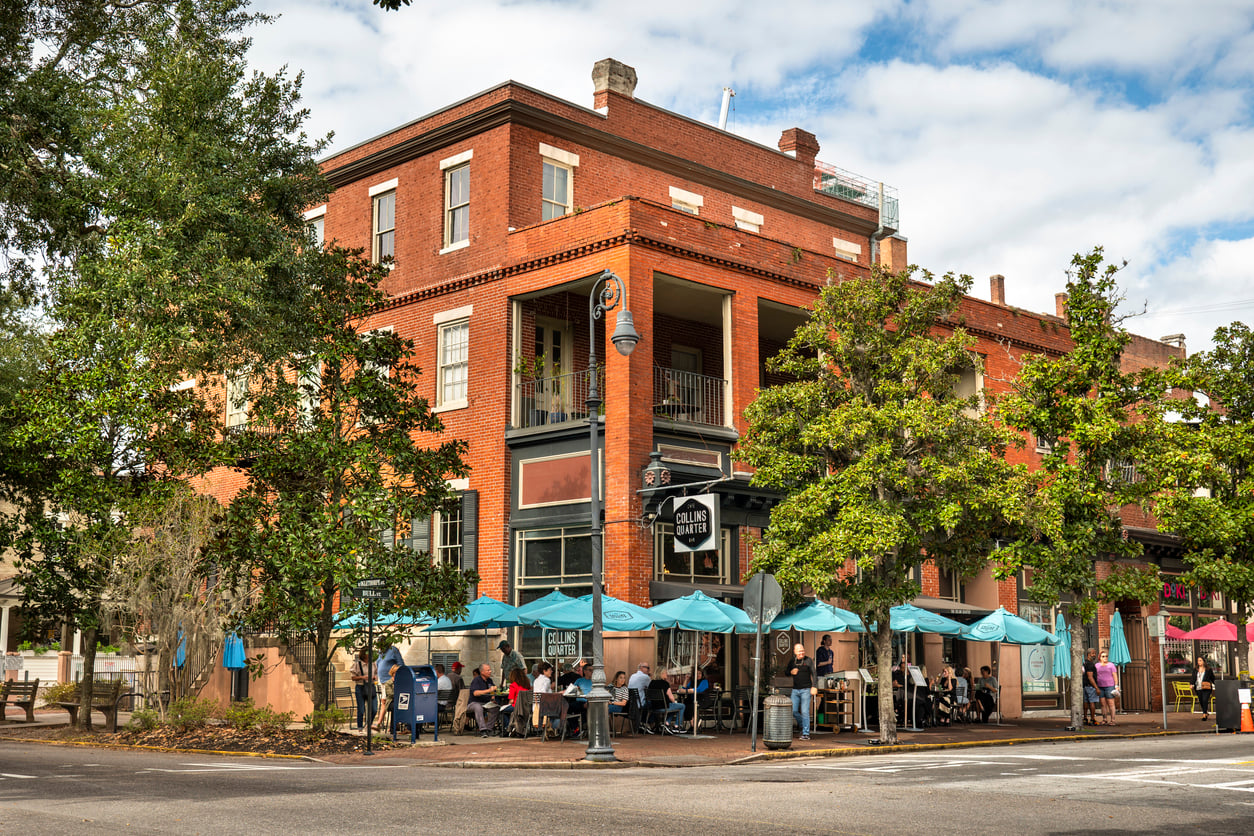 A corner brick building houses a café with outdoor seating under blue umbrellas; people are dining, and leafy trees line the sidewalk on a partly cloudy day.