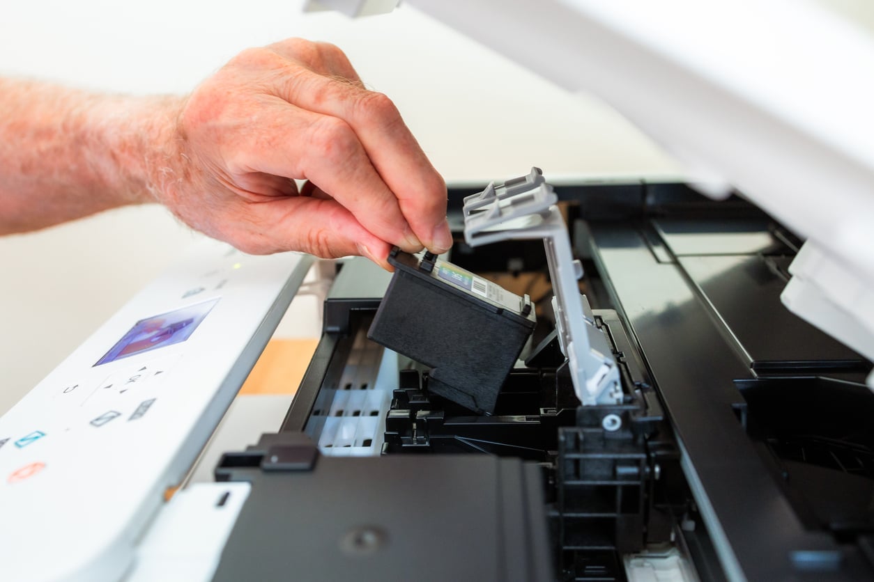 A person’s hand inserts or removes a black ink cartridge inside an open inkjet printer—a process familiar to many who have experienced waste of money products—with the printer cover lifted and printer controls visible on the left.