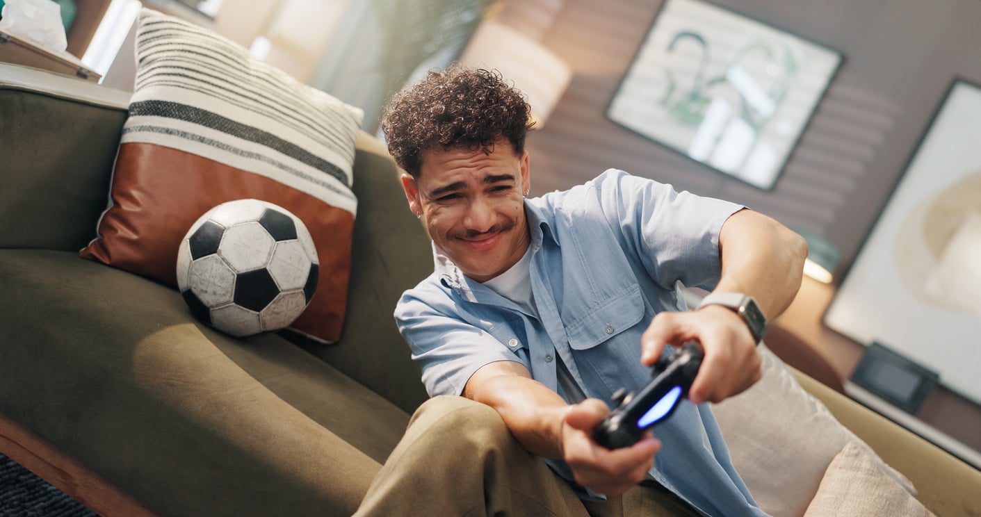 A young man sitting on a couch, holding a game controller with a focused expression, indulges in one of his expensive hobbies. A soccer ball and a striped pillow are beside him, and framed pictures hang on the wall in the background.