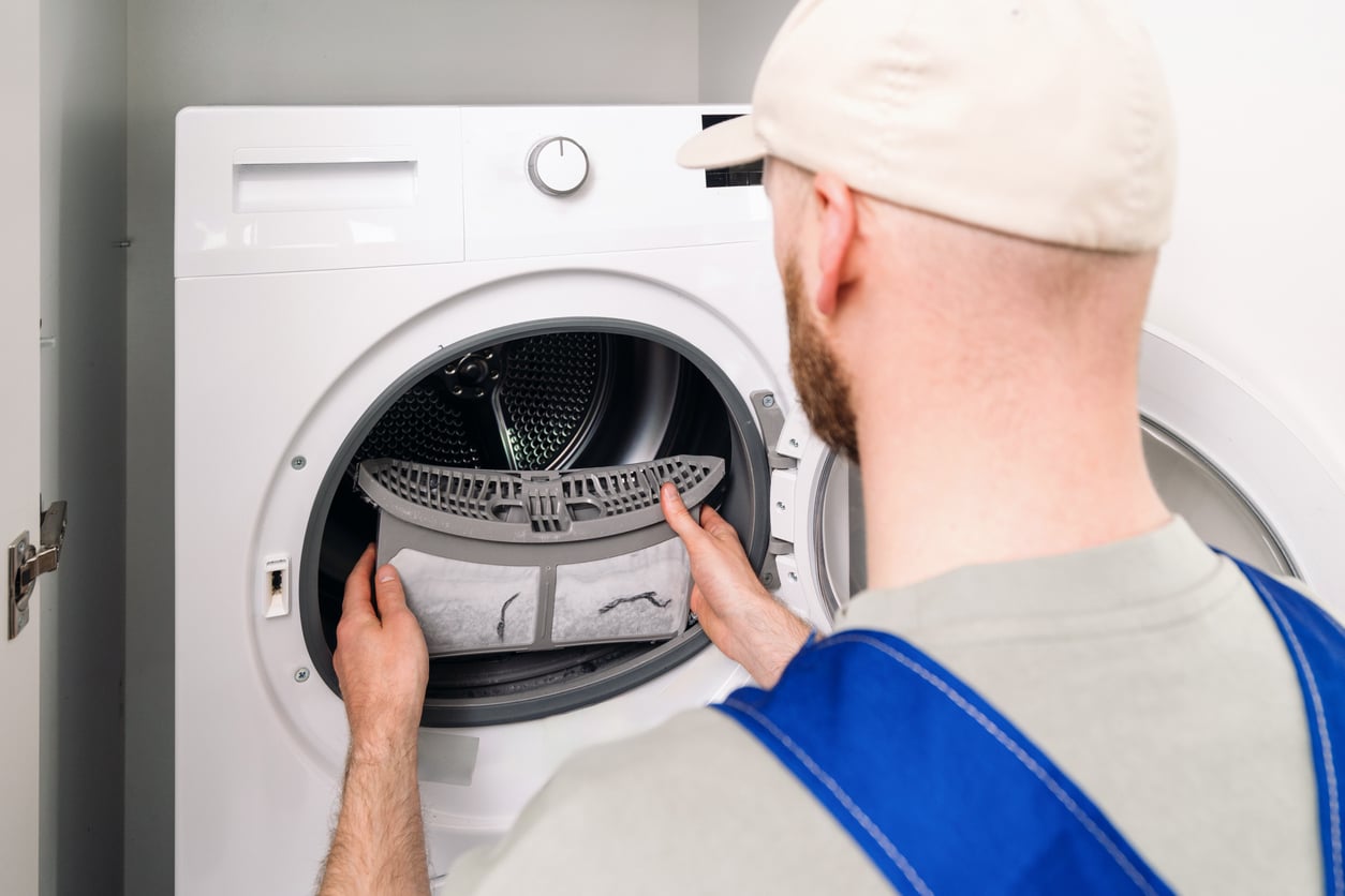 A person in a cap and work overalls cleans the lint filter of a front-loading clothes dryer, holding the filter in both hands near the open door of the appliance.